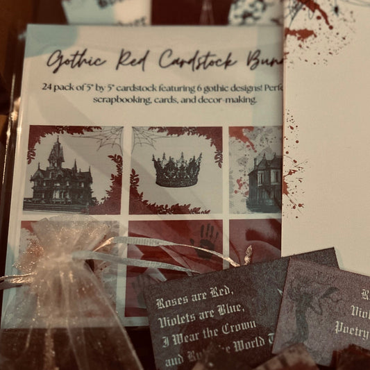 A boxed set of Gothic Red cardstock bundles and scrapbooking supplies arranged inside a cardboard package. Visible items include printed cardstock sheets with gothic house illustrations, small cards, a clear bag with silver chains, and a small wrapped accessory, all themed around a dark red and cream color story.